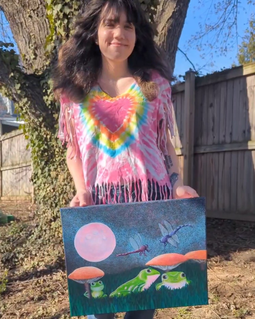 Photograph of Heather Thomas, a brunette woman standing in front of a tree holding a painting